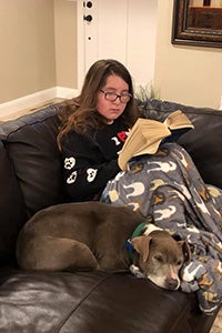 Gianna sitting on a couch reading with a blanket over her lap and Yvonne the dog sleeping next to her