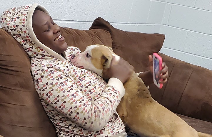 Woman sitting on a couch wearing a hoodie with a brown and white dog leaning up for some love