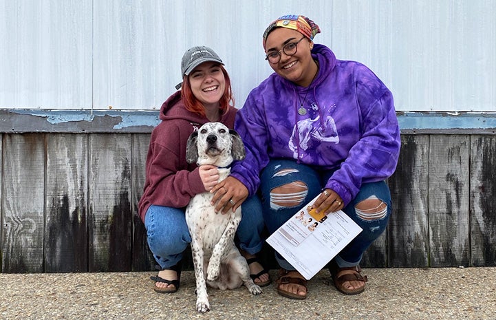 Two women squatting down next to a wall hugging a white dog with black spots