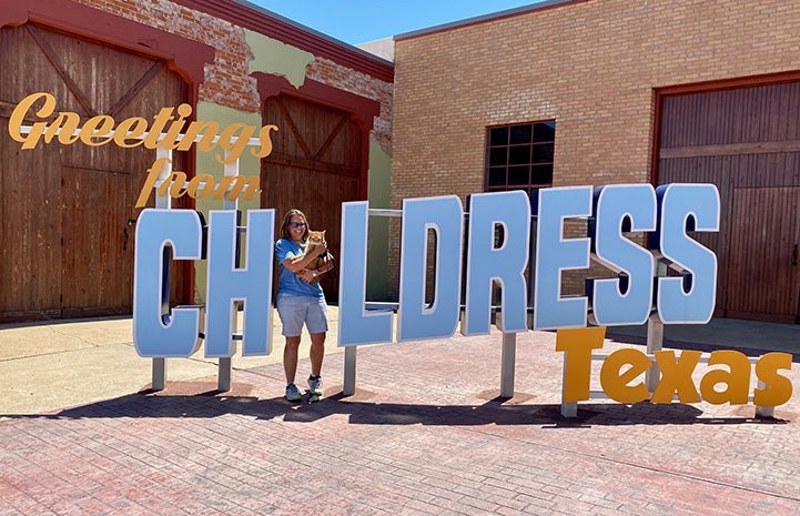 Woman holding Steak the cat in front of a Greetings from Childress, Texas sign