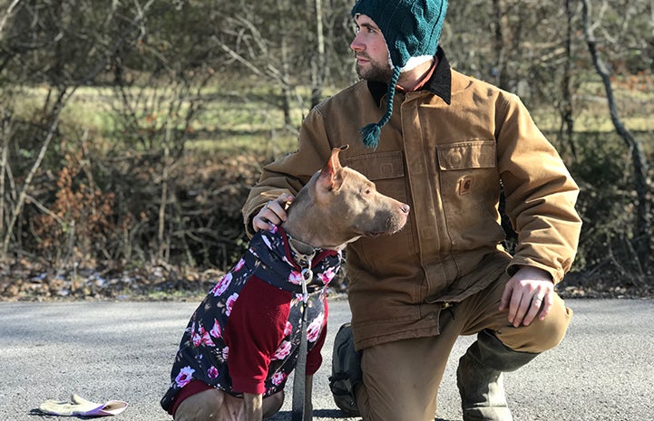 Man bundled up in winter weather protective clothing next to Kiwi the dog in a sweater