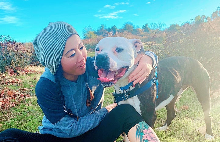 Woman wearing a knit hat and sitting in a meadow with Zeke the dog