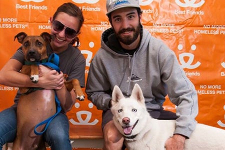 Ashley and Travis Tzioumis with two dogs in front of a Best Friends backdrop