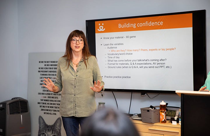 Sarah Hock addressing an audience with a PowerPoint presentation on a screen behind her