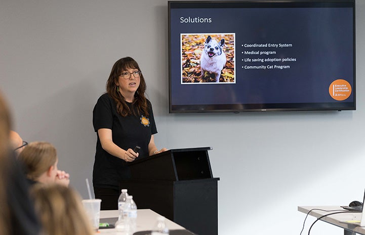 Sarah Hock at a podium addressing a class with a Solutions PowerPoint slide on a screen behind her