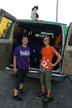 Two women standing at the back of a van, with its doors open and a cat on top