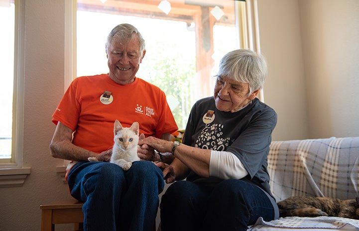 Narnia the flamepoint Siamese cat sitting on the lap of Vann, who is sitting on a couch next to his wife Mary Rose