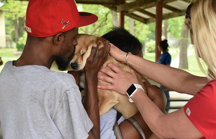 Man wearing a red hat kissing the head of a small brown dog