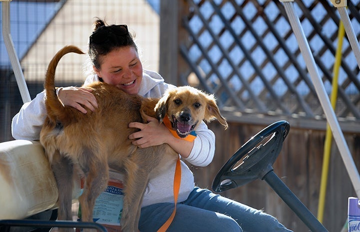 Woman sitting in a golf cart hugging a smiling brown dog