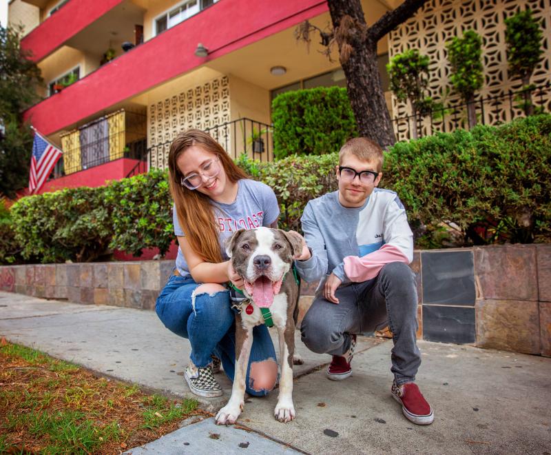 A couple and happy pit bull type dog outside in front of a home