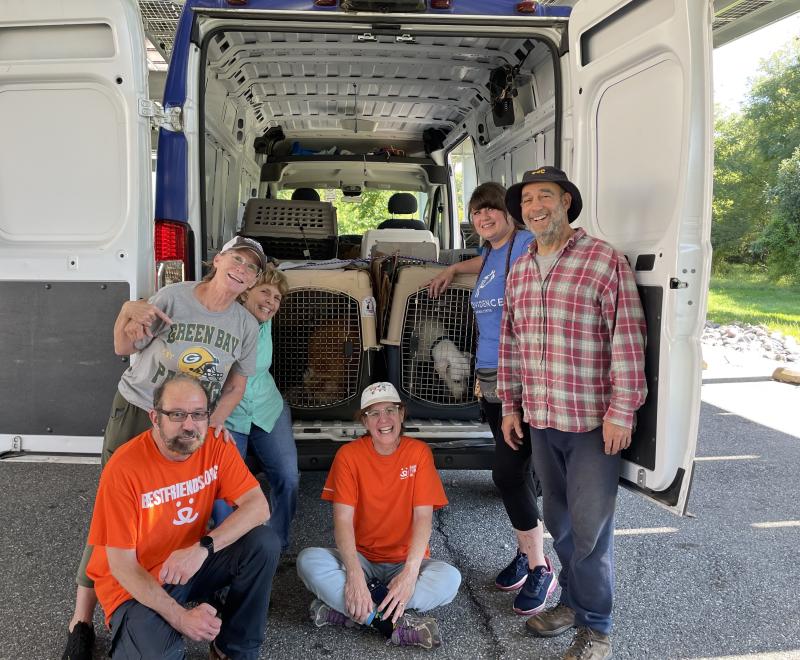 Six smiling people at the back of a van with the back doors open, showing multiple animal kennels