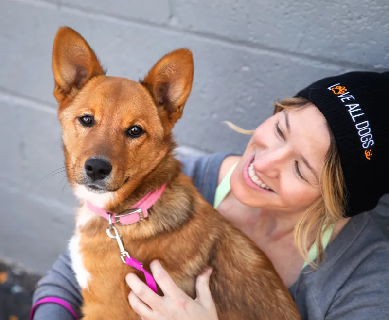 Smiling person sitting with dog outside next to a gray wall