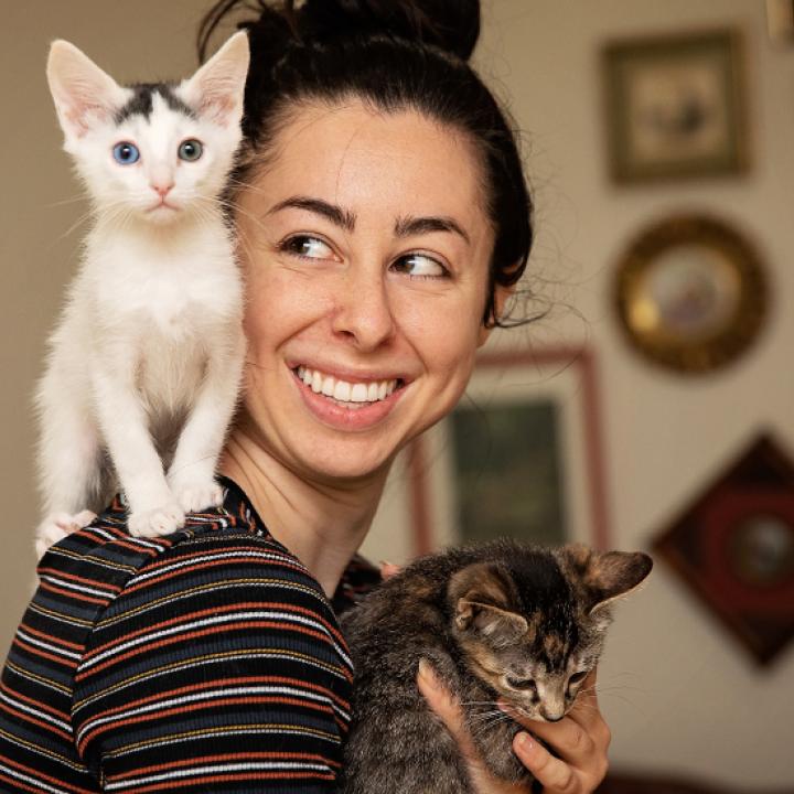 Smiling person holding a kitten with another black and white kitten on her shoulder