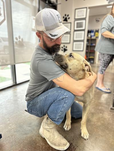 Person wearing a hat and kneeling down and hugging a shepherd mix dog