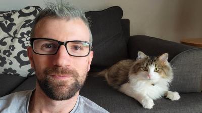 Selfie of Scott in the foreground with Dolly Parton the cat lying on the couch behind him