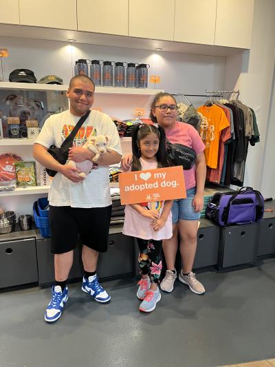 Family of three with one holding a small white puppy and another holds an "I heart my adopted dog" sign at the Best Friends Lifesaving Center in New York City