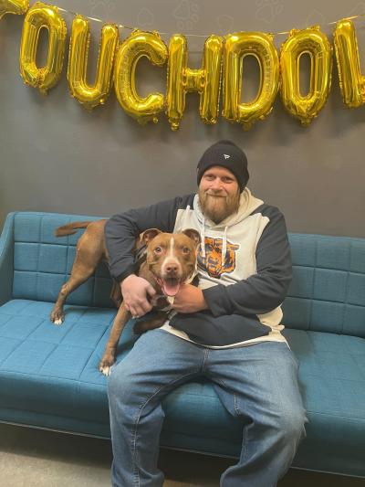 Person sitting on a blue couch with a happy brown dog under balloons spelling the word Touchdown