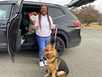 Smiling person in front of a vehicle with a rear door open with a shepherd dog on a pink leash