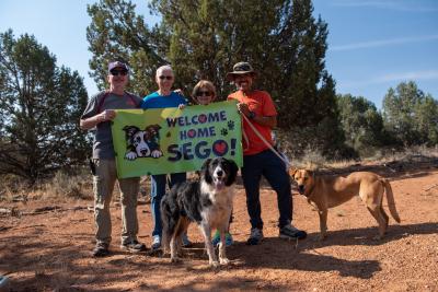 People outside holding a banner that says "Welcome Home Sego" with two dogs including Sego