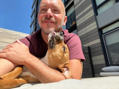 Volunteer David Sprague cradling a brown and white dog in his arm and lap, who is licking David's chin