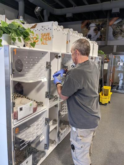 Jeff Harris the volunteer wearing rubber gloves, holding a bowl in front of some white cat kennels