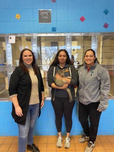 Three people standing in front of cat kennels, with one person wearing a Best Friends sweatshirt holding a tabby cat