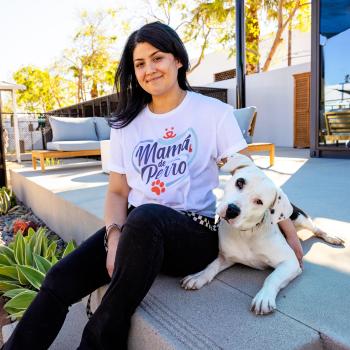 Smiling person wearing a shirt that says "Mamá de Perro" sitting outside on some with a big dog next to them